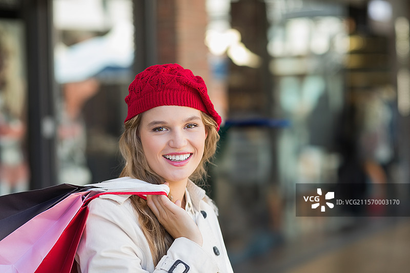 微笑的女性在购物区提着五颜六色的纸购物袋，戴着红色针织帽。图片素材