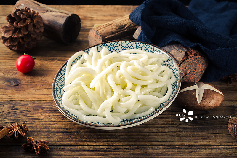青花瓷碗中盛放洁白弹滑的年糕条，造型饱满诱人。背景为古朴木桌、松果香料与香菇原木，整体氛围温馨质朴图片素材