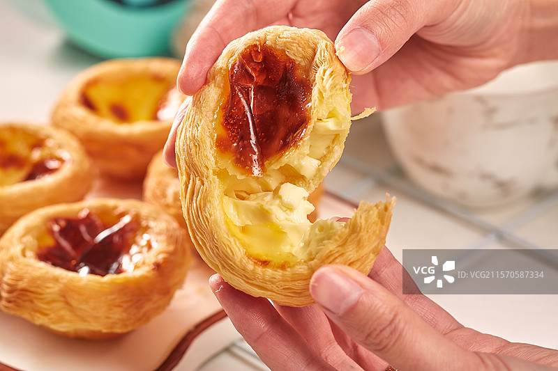 双手正掰开香喷喷的一个蛋挞且桌上还放着几个完整的蛋挞图片素材