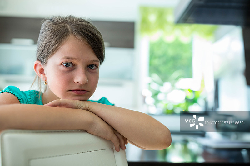 女孩穿着青绿色上衣，靠在厨房的白色椅子上，窗外模糊的树叶映入眼帘。图片素材