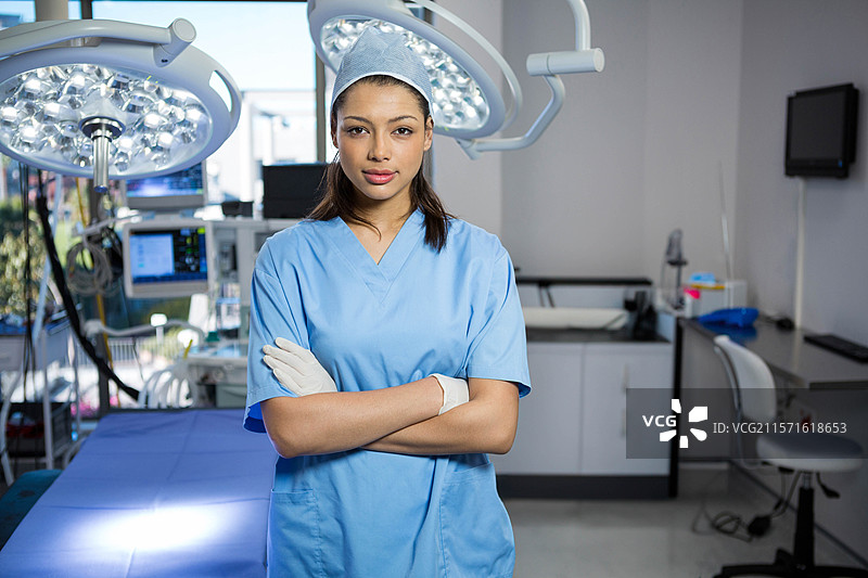 穿着蓝色手术服的女性外科医生双臂交叉，站在LED灯和监视器下的手术室内。图片素材