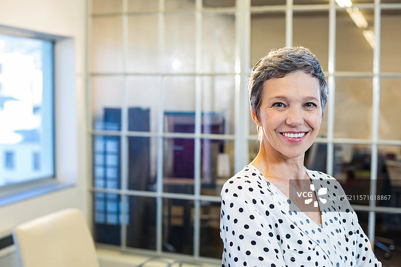 一位年长女性专业人士微笑着站在办公室内，双臂交叉，靠近玻璃隔断墙。图片素材