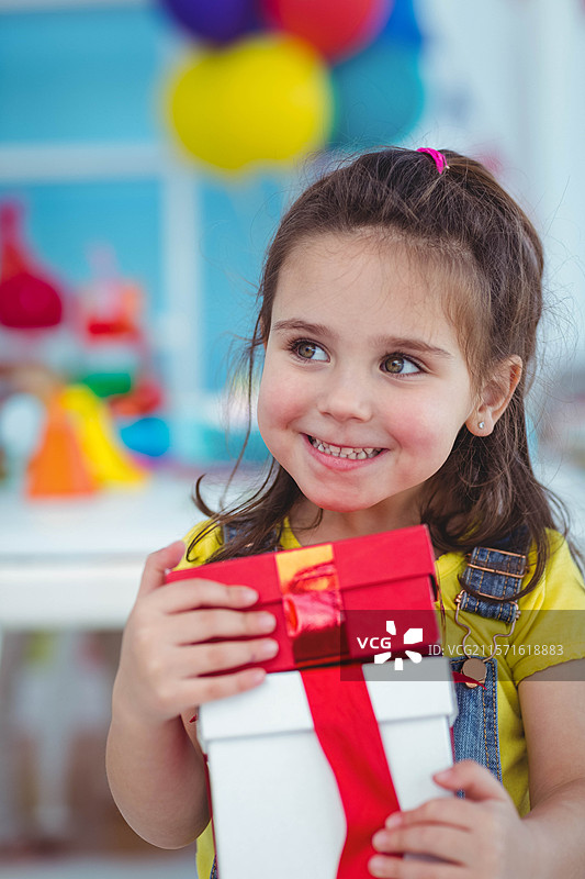幼儿园女孩在派对房间里手持两层礼品盒，朝右侧看，周围是气球和玩具。图片素材