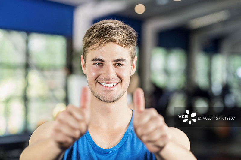 穿着蓝色背心的男子微笑着，竖起大拇指，站在健身房的器械旁。图片素材