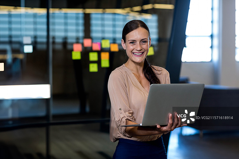 女性在现代办公室会议区微笑着手持银色笔记本电脑，周围有霓虹色的便签。图片素材