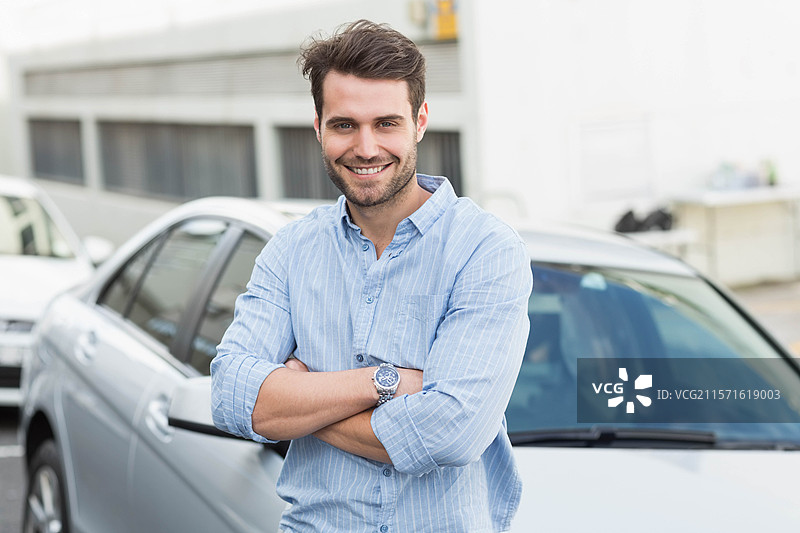 男子双臂交叉，佩戴手表，站在汽车经销商处，旁边是一辆银色轿车。图片素材