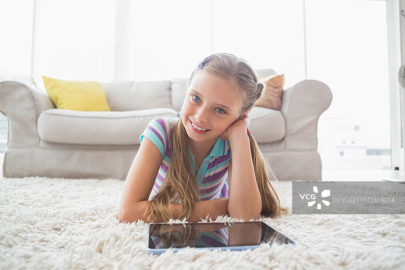 学龄女孩躺在明亮的客厅里，柔软的白色地毯上，凝视着平板电脑。图片素材