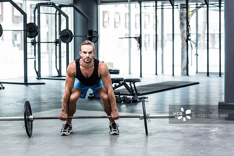 男性举重运动员在健身房的台子上进行硬拉，周围有深蹲架、卧推架和哑铃组。图片素材