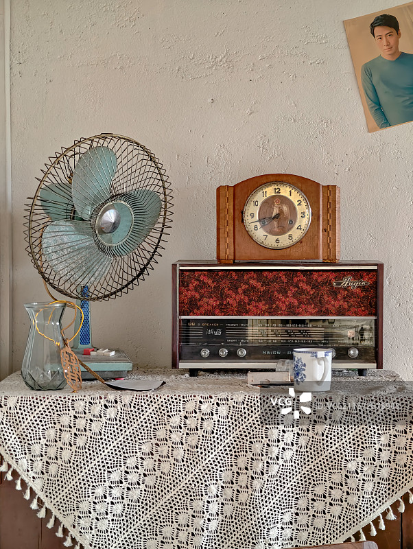 20世纪80年代房间:白色钩花桌布、老式电风扇、复古座钟、木质收音机、玻璃花瓶与青花瓷杯以及明星海报图片素材