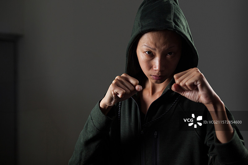 中年亚洲女性在昏暗的训练室里举起拳头，穿着绿色连帽卫衣。图片素材