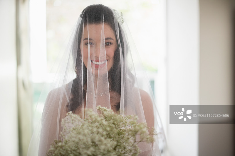 新娘站在明亮的入口处，戴着面纱，穿着无肩带礼服，手捧满天星花束。图片素材