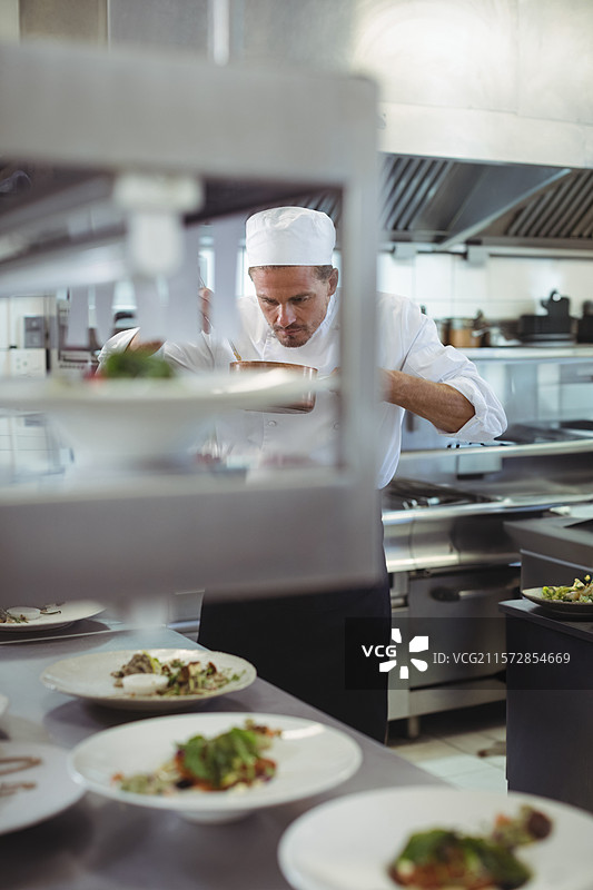 男性厨师俯身在台面上为瓷盘上的菜肴装饰配料，放在厨房的传递架上。图片素材