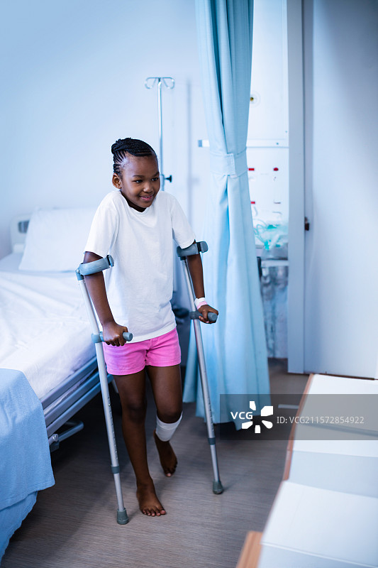 在医院里，一位非裔美国女孩用拐杖支撑着包扎的膝盖，旁边有输液架。图片素材