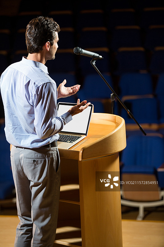 演讲者在空荡荡的蓝色椅子前，站在木质讲台后，手势表达，讲台上有笔记本电脑和麦克风。图片素材