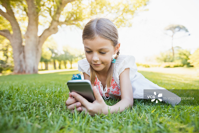 阳光明媚的公园里，穿着红色刺绣裙子的学龄女孩躺在绿色草坪上，手里拿着智能手机。图片素材