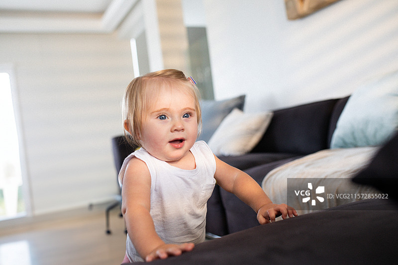 女婴站着，靠在深灰色沙发垫上，身处窗帘旁的客厅。图片素材