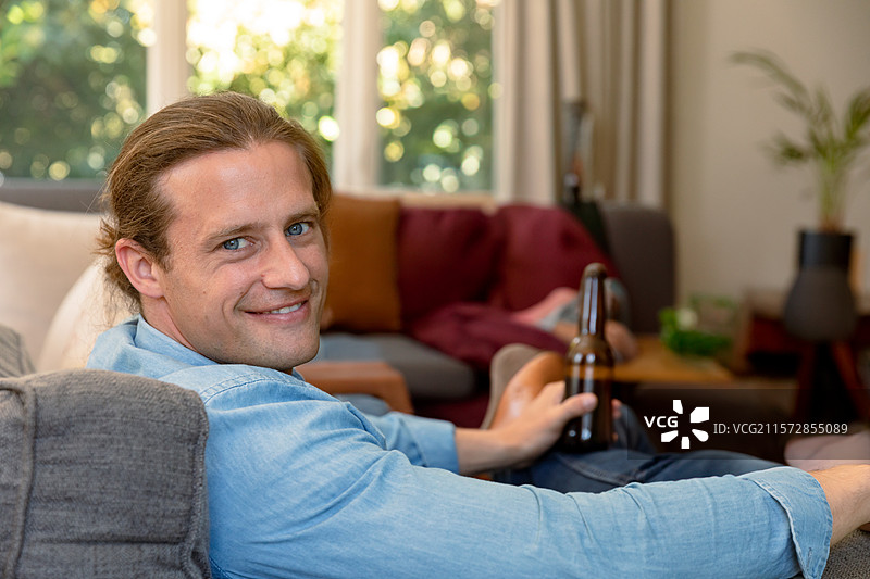 男性靠在灰色沙发上，手里拿着深色啤酒瓶，旁边有一盆植物。图片素材