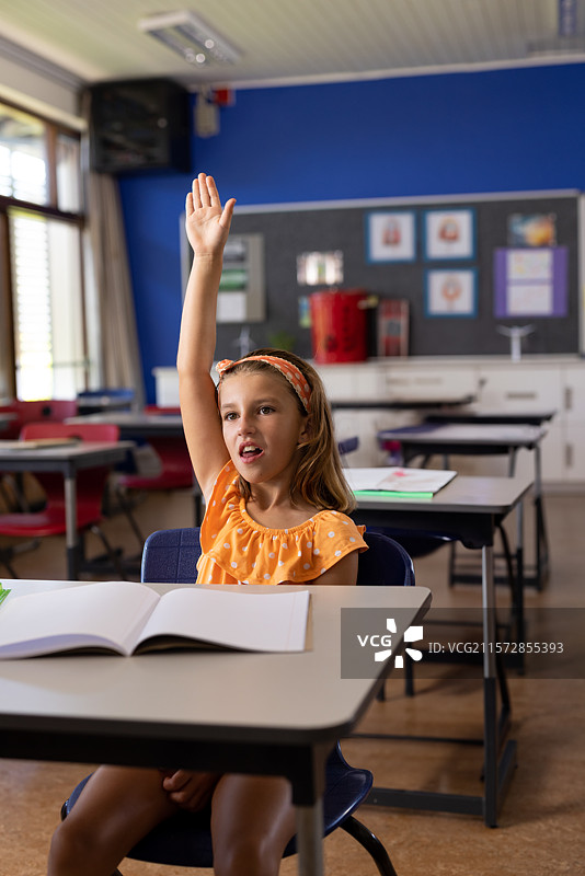 小学女孩在教室的桌子上举手，手里拿着铅笔，旁边有一本打开的笔记本。图片素材