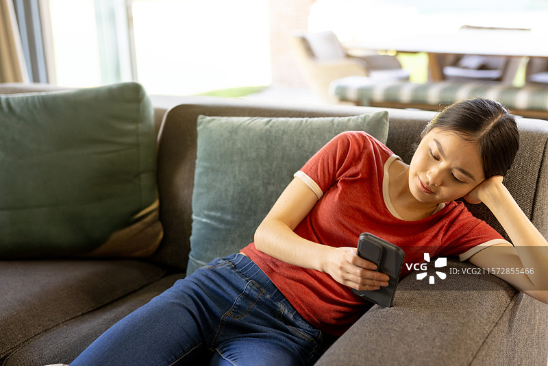 亚洲女性在客厅的灰色沙发上靠着，手持智能手机，留有空白空间图片素材