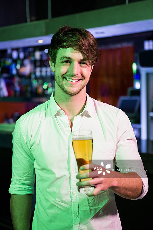 男子在酒吧柜台下的绿色灯光下举着一杯啤酒，展示着酒瓶和收银机。图片素材