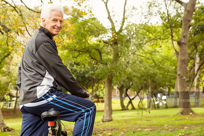 穿着拉链夹克和运动裤的老年男子微笑着骑自行车穿过公园，留有空白空间图片素材