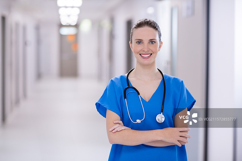 穿着蓝色护士服的女性医护人员站在医院走廊，手中拿着听诊器。图片素材