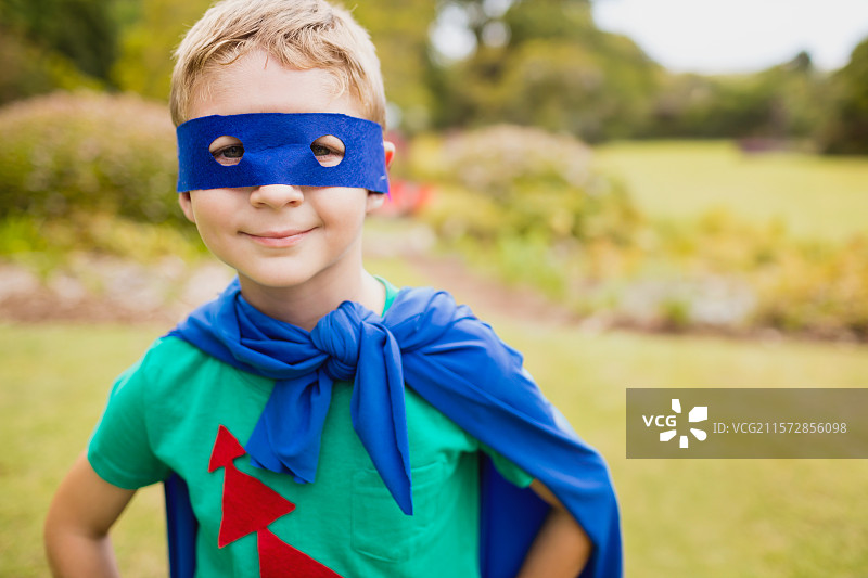 男孩在花园中摆姿势，双手叉腰，戴着蓝色毛毡面具和配套斗篷。图片素材
