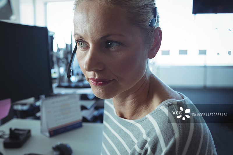 在开放式办公室的桌子前，成年女性佩戴耳机接听电话并查看电脑显示器。图片素材