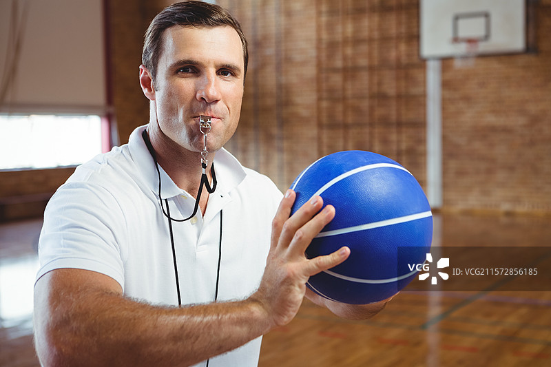 成熟的成年男性裁判手持蓝色篮球，吹着哨子站在体育馆的场地上图片素材