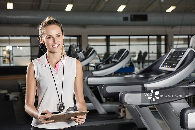 女性健身教练微笑着站在健身房的跑步机旁，手持夹板和笔，佩戴计时器。图片素材