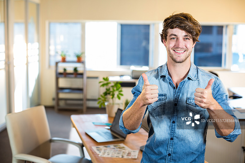 穿着牛仔衬衫的男人在木桌后竖起大拇指，桌上有笔记本电脑和植物，留有空白空间图片素材
