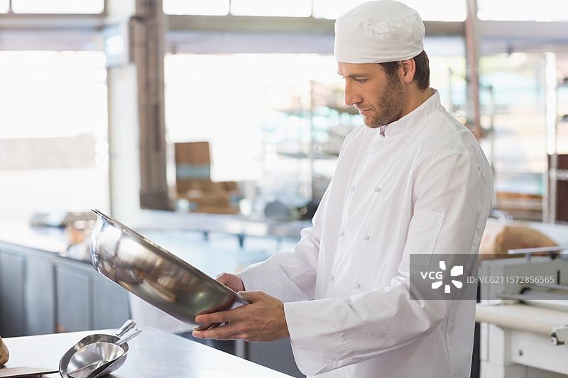 穿着制服的男厨师在面包店厨房里手持不锈钢搅拌碗进行检查，留有空白空间图片素材