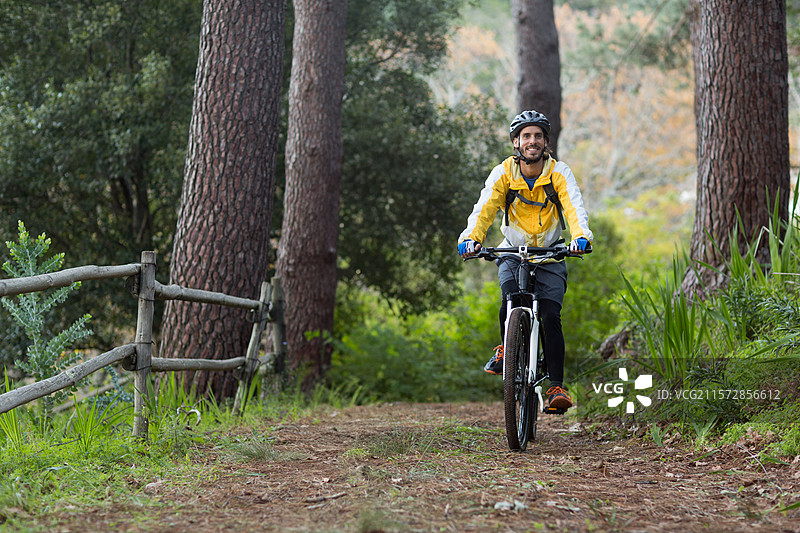 男性骑自行车者佩戴黄色头盔，背着背包，在森林小径上沿着木栅栏骑行。图片素材