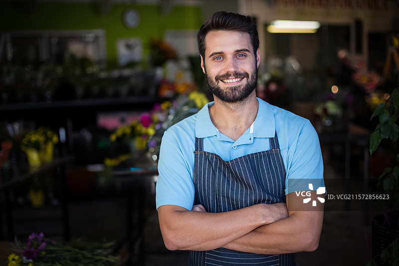 男子双臂交叉站在花店的木质展示桌旁，正在整理五彩缤纷的花束。图片素材