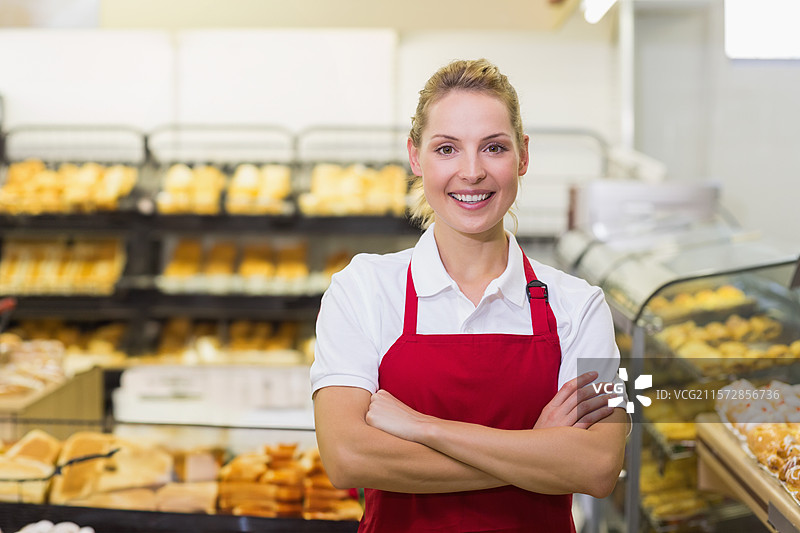 二十多岁的女性穿着红色围裙，站在面包店的柜台后面，旁边有面包架和玻璃展示柜。图片素材