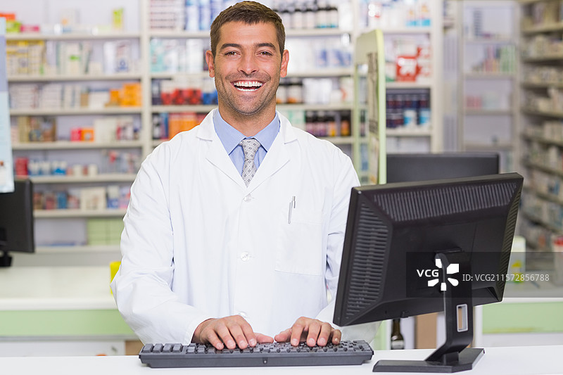 男性药剂师站在药房柜台前，正在键盘上输入，身后是药品货架。图片素材