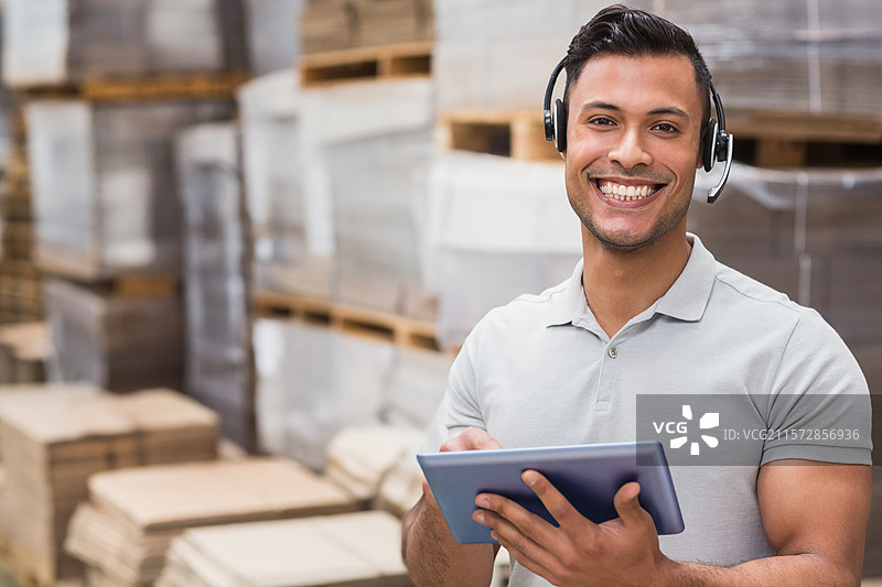 穿着灰色 polo 衫的成年男性仓库工人戴着耳机，站在箱子旁边，手持平板电脑，留有空白空间。图片素材