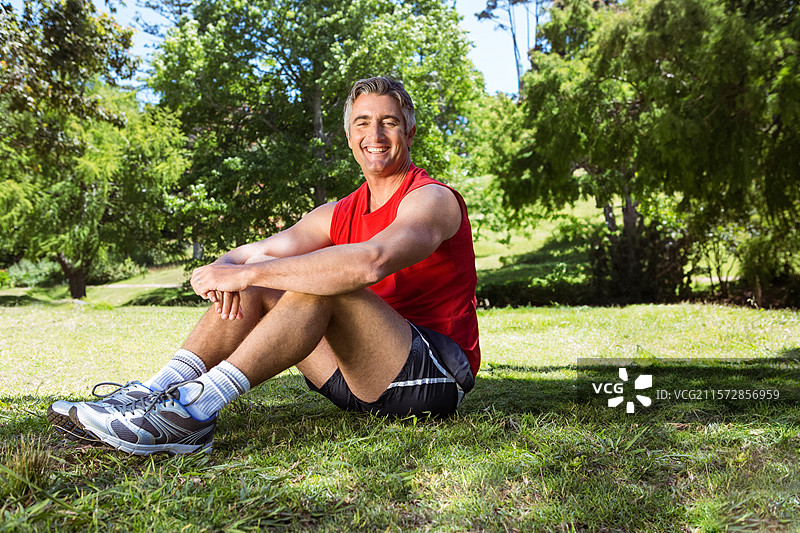 一位老年男子坐在公园阳光明媚的草地上，双膝弯曲，微笑着，穿着灰色跑鞋。图片素材