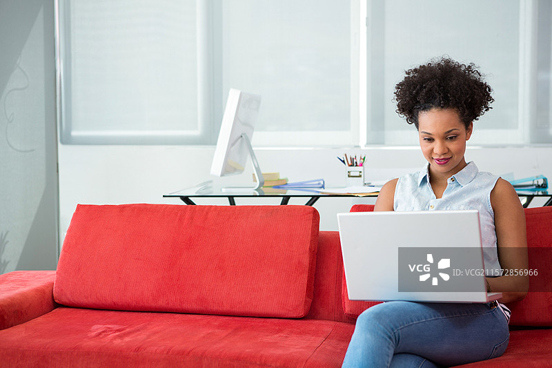 非裔美国女性在办公室休息室的红色沙发上使用笔记本电脑，旁边有显示器，留有空白空间。图片素材