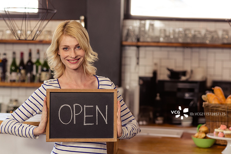 穿着条纹上衣的女性在咖啡机和糕点的柜台上举着“开放”黑板。图片素材