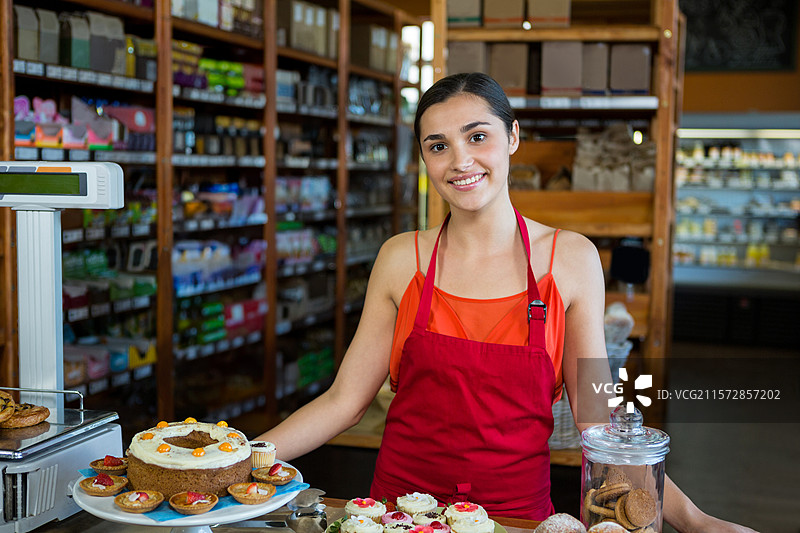 穿着红色围裙的女人在柜台后微笑，柜台上展示着裱花蛋糕、杯子蛋糕和饼干罐。图片素材