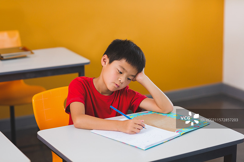 亚洲男孩用手托着头，在学校的课桌上用铅笔在五彩的作业本上写字。图片素材