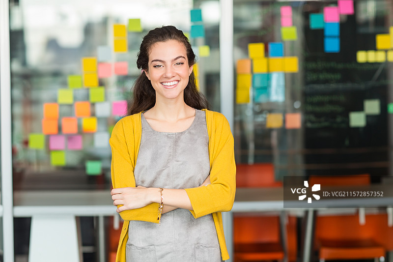 女性专业人士微笑着站在玻璃墙旁，会议桌旁贴满了便利贴。图片素材