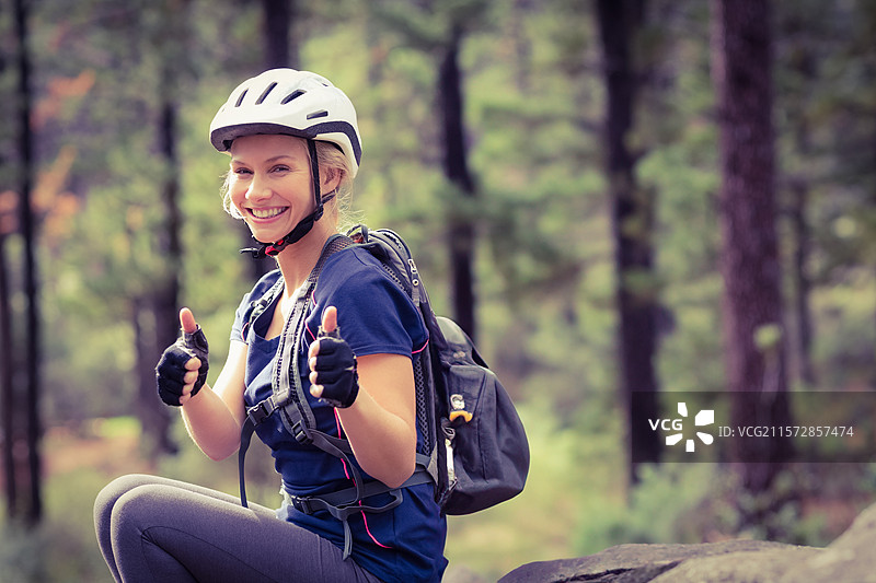 穿着白色自行车头盔和手套，背着黑色背包的女人坐在松树林中的岩石上图片素材