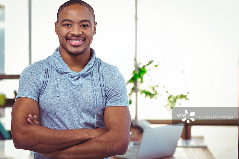 站着的男人双臂交叉，微笑着站在展示笔记本电脑和植物的桌子旁，窗户附近。图片素材
