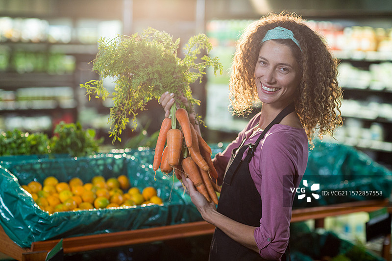 女性蔬菜专员手持胡萝卜，在橙色箱子旁整理货架，留有空白空间图片素材