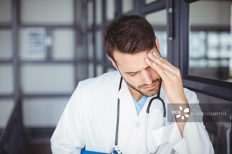 男医生靠在医疗走廊的玻璃柜上，手里拿着蓝色文件夹和听诊器。图片素材