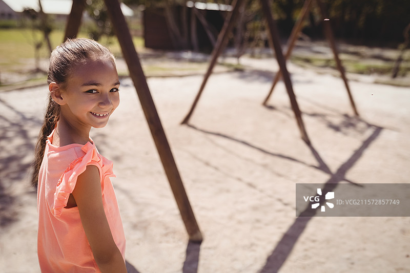 在郊区公园的沙地游乐场上，站着一个上学年龄的女孩，旁边有木制秋千架，留有空白空间。图片素材