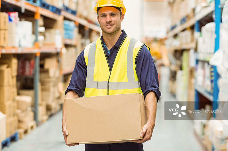 男性仓库工人戴着安全帽和背心，手里拿着纸箱，站在金属货架之间的过道上。图片素材
