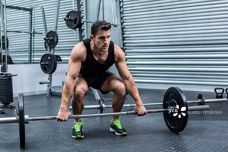 在健身房里，穿着绿色鞋带黑色鞋子的运动男士正在深蹲，旁边是杠铃架。图片素材